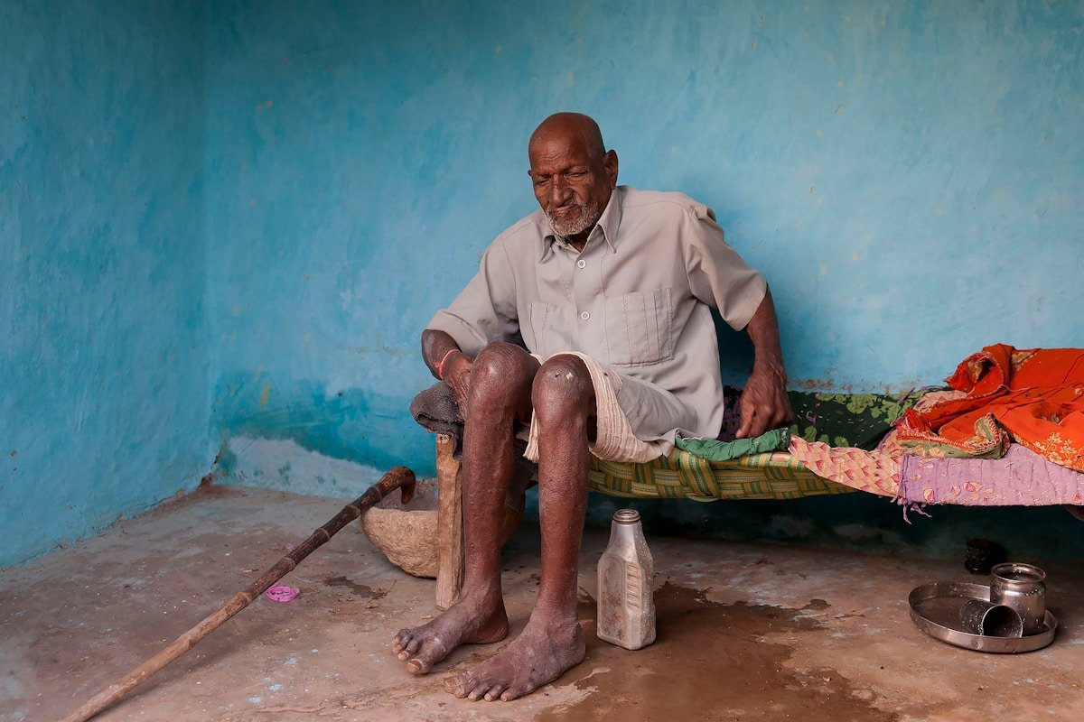 Homme âgé, assis, peau foncée, vêtements usagés, environnement modeste.